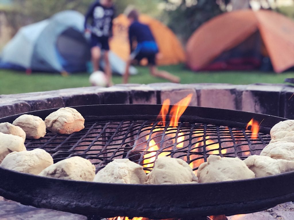 Making roosterkoeke dough bread on a grid on a wood fire in a fire pit selective focus with two boys playing football soccer in front of tents in South Africa