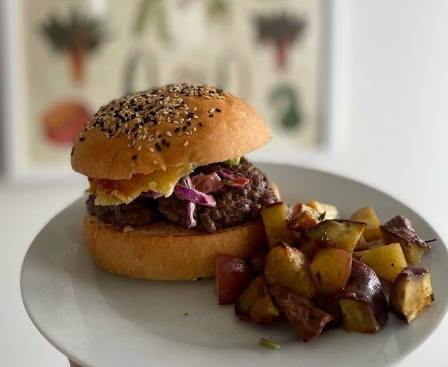 Homemade burger on a white plate with roasted kumara, held in front of a vegetable poster