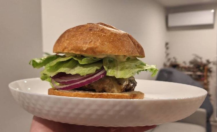 Profile of Crumbed Chicken burger on a focaccia bun with lettuce, red onion and cheese, on a white plate held by hand
