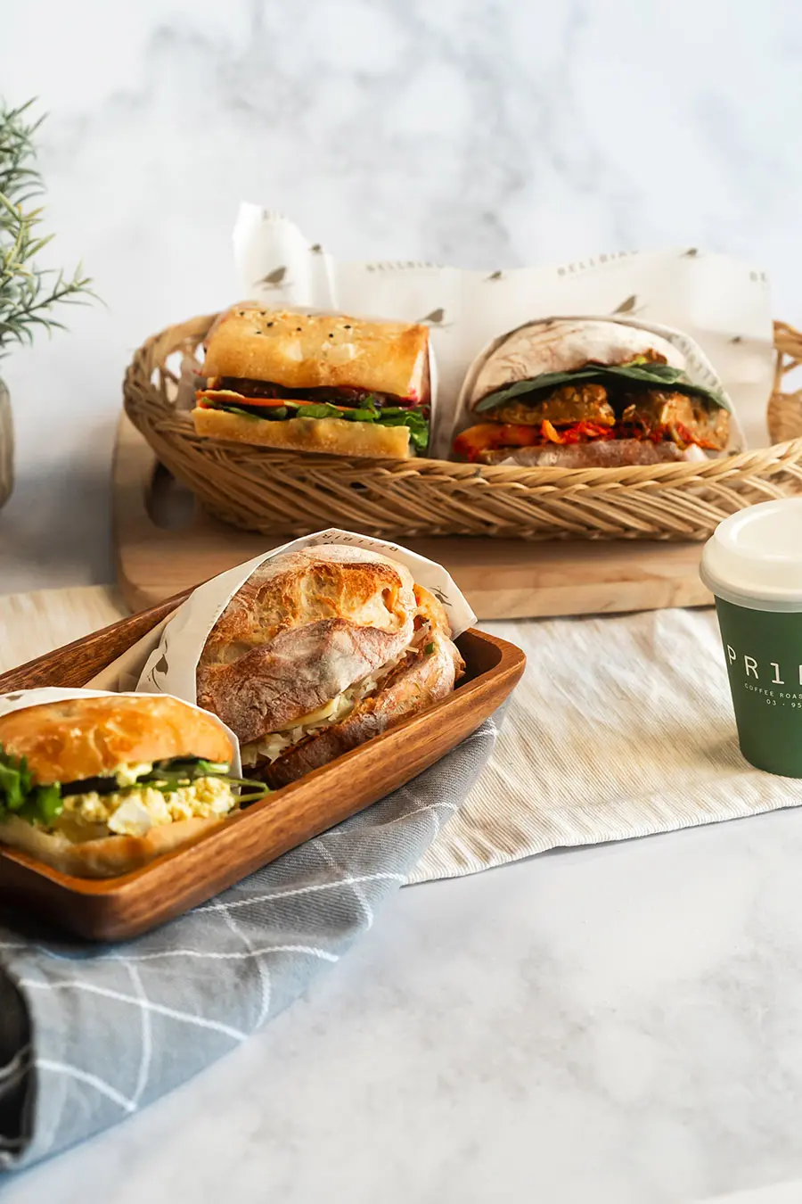 Range of different sandwiches from Bellbird Bakery, Egg, Reuben, Tempeh, Kimchi. On wood board and in baskets with Coffee Cup