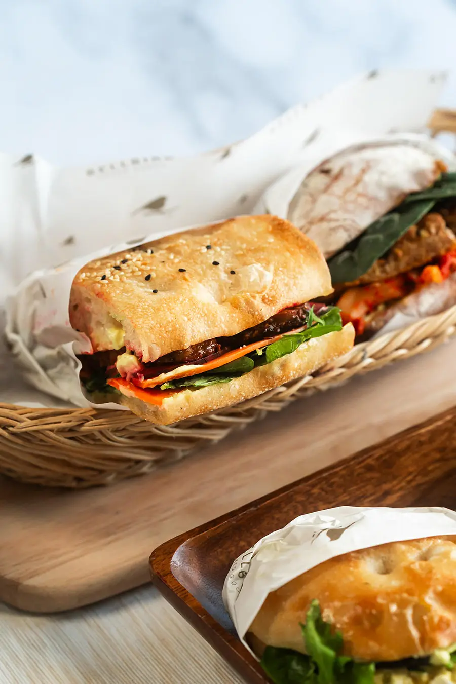 Tempeh Sandwich wrapped and in wood basket next to Kimchi and Egg Sandwich