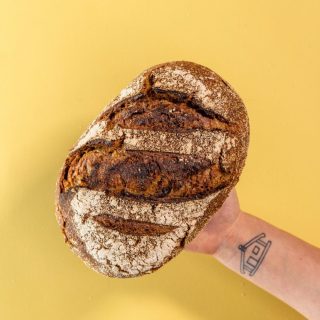 Spelt Sourdough loaf held by tatooed arm on yellow background